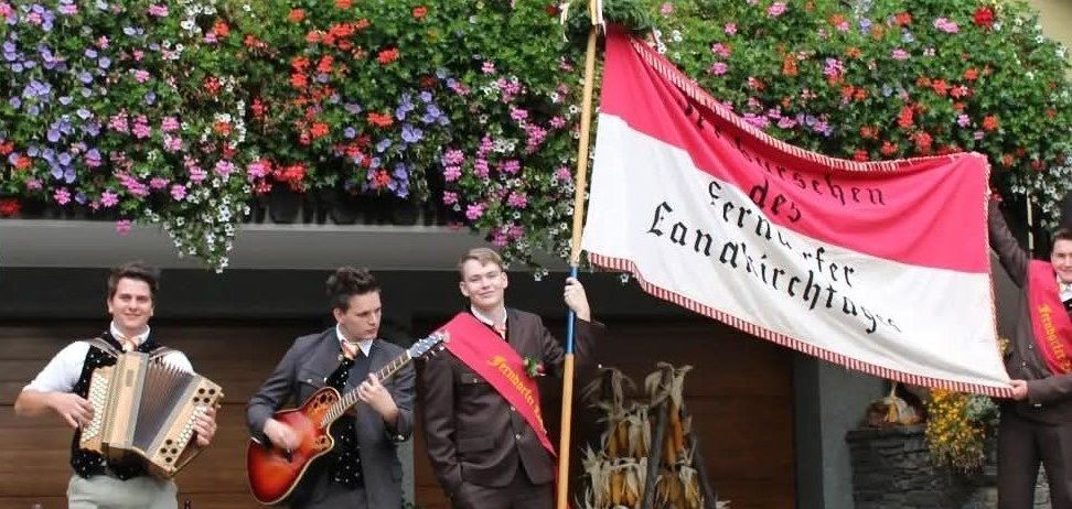 Vier Männer in Anzügen und Krawatten spielen Instrumente draußen und halten eine Fahne mit der Aufschrift 'Erntedankfest'. Hinter ihnen befindet sich ein Gebäude, das mit Blumen geschmückt ist.