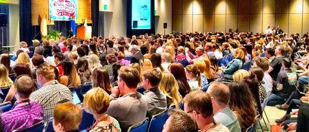 A large audience in a conference room listens to a presentation on a large screen. The attendees, seated in blue chairs, are engaged and using their laptops. The presenter stands on stage with a microphone, and the room is well-lit with a yellow ceiling.