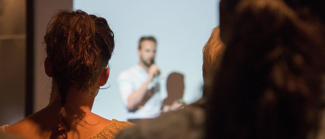 Ein Mann hält eine Präsentation mit einer Projektionsleinwand im Hintergrund. Die Leute sehen von vorne zu.