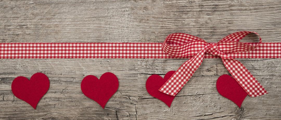 Eine hölzerne Oberfläche mit einem roten Gingham-Band und vier roten herzförmigen Objekten. Das Band ist zu einer Schleife gebunden und hat ein kariertes Muster. Die Herzen sind auf dem Band platziert.