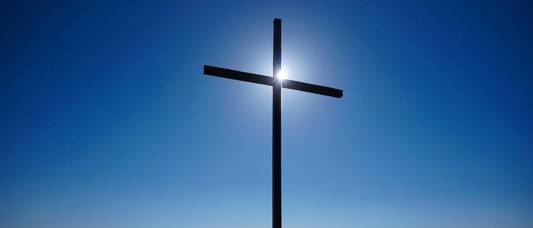 Ein großes Kreuz steht auf einem Hügel, die Sonne scheint hell durch es gegen einen blauen Himmel und überblickt eine weite grüne Landschaft.