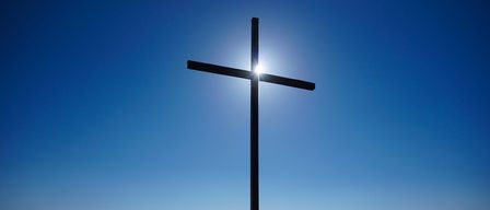 Ein großes Kreuz steht auf einem Hügel, die Sonne scheint hell durch es gegen einen blauen Himmel und überblickt eine weite grüne Landschaft.