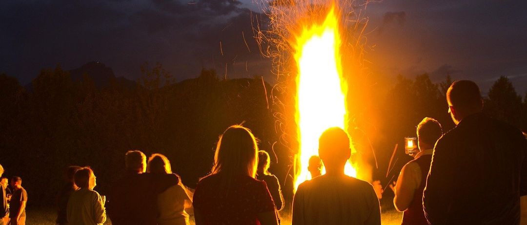 Ein Lagerfeuer brennt hell im Freien bei Nacht, mit Menschen, die herumstehen und einige Fotos machen. Der Himmel ist teilweise bewölkt.