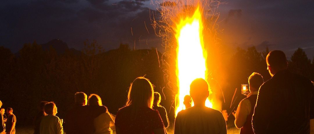 Eine Gruppe von Menschen versammelt sich bei Nacht um ein großes Lagerfeuer, einige halten Handys, um den Moment festzuhalten. Das Feuer wirft Funken, und der Himmel ist dunkel mit verstreuten Wolken.