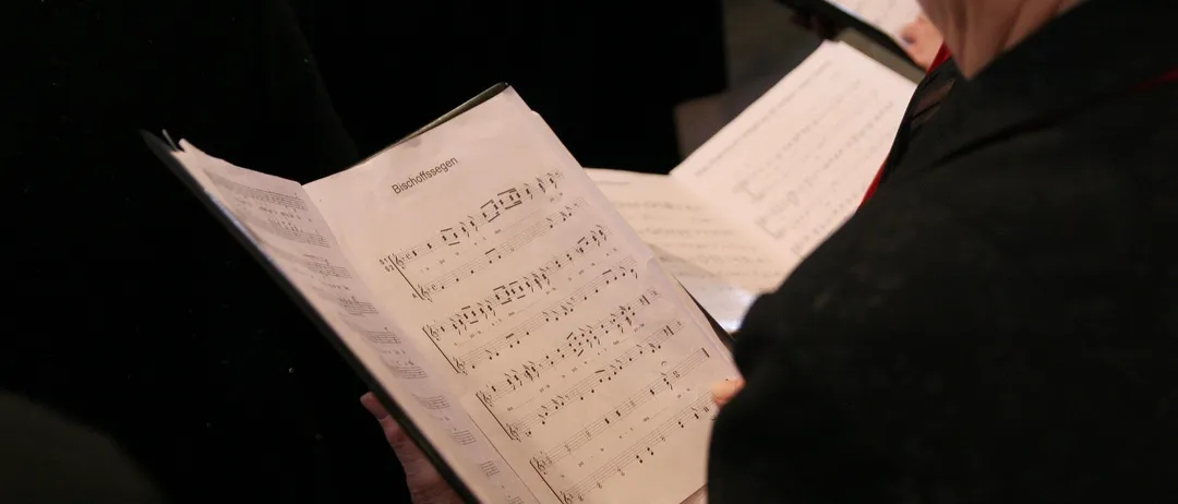 Eine Person hält ein offenes Notenbuch mit Musiknoten, wahrscheinlich während einer Chorprobe. Der Hintergrund ist unscharf mit einer anderen Person, die ein Musikbuch hält.