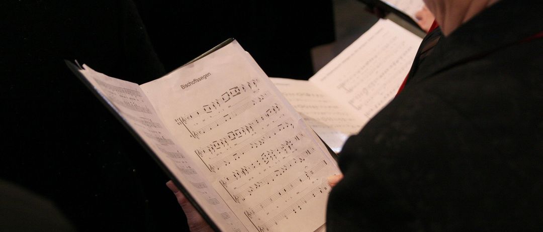 Eine Person hält ein offenes Notenbuch mit Musiknoten, wahrscheinlich während einer Chorprobe. Der Hintergrund ist unscharf mit einer anderen Person, die ein Musikbuch hält.