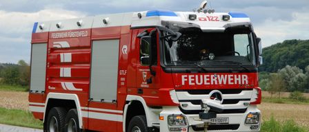 Ein roter und weißer Feuerwehrwagen mit der Aufschrift 'FEUERWEHR' auf der Vorderseite steht am Straßenrand.