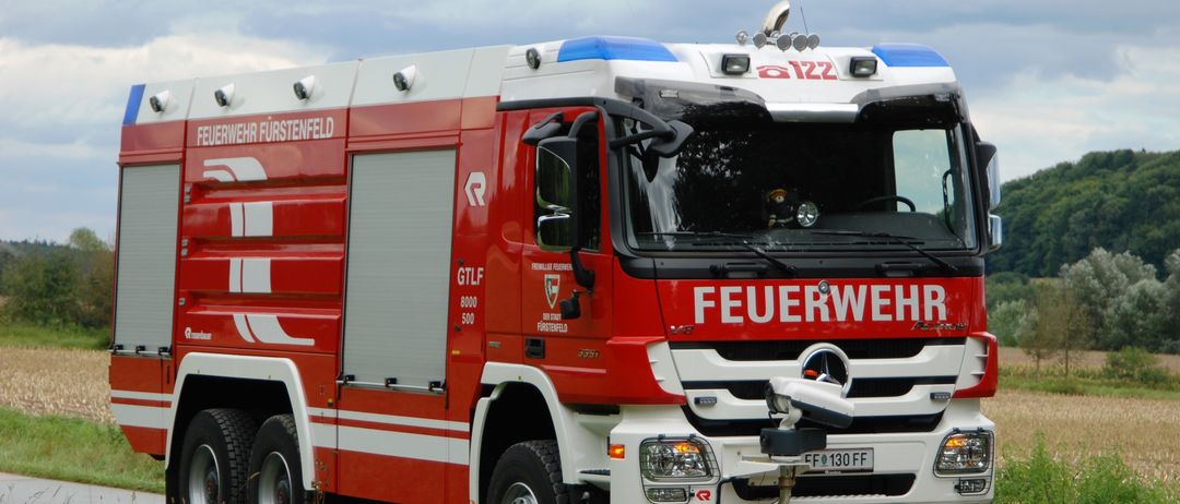 Ein roter und weißer Feuerwehrwagen mit der Aufschrift 'FEUERWEHR' auf der Vorderseite steht am Straßenrand.