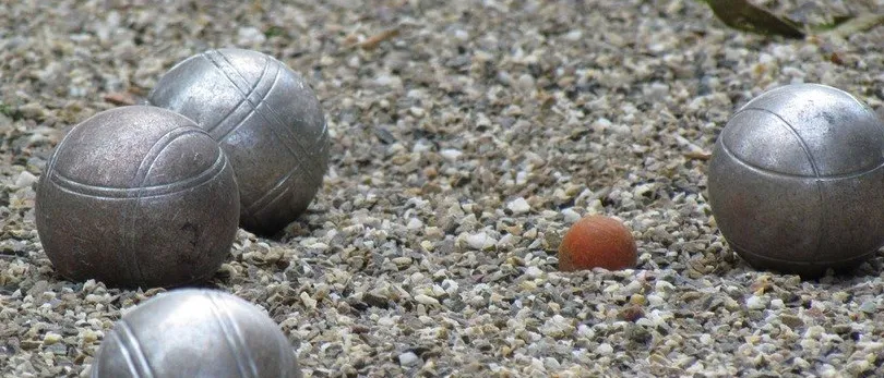 Vier metallische Bälle, drei größere und einer kleinere, liegen auf einer Kiesoberfläche. Die größeren Bälle sind grau, während der kleinere rötlich-braun ist.
