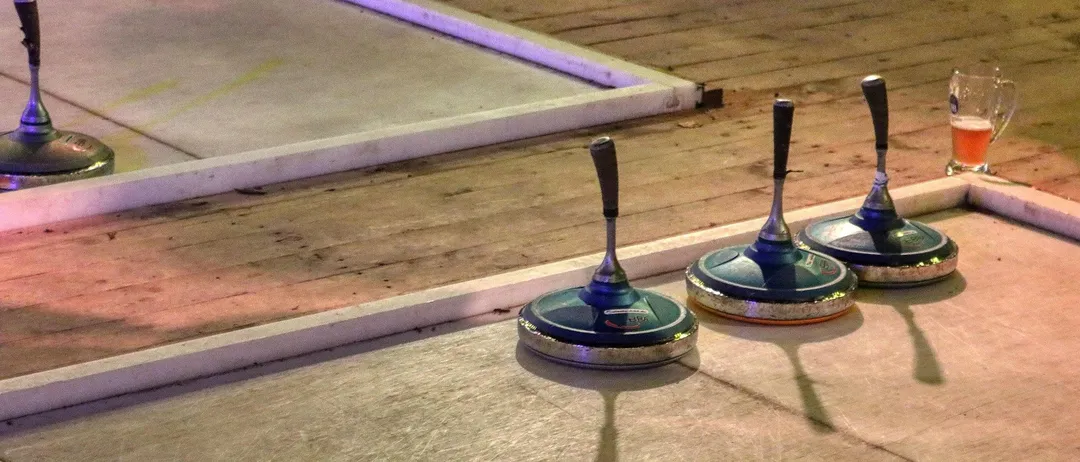Drei Curlingsteine sind auf einem polierten Boden platziert, wobei Schatten auf der Oberfläche geworfen werden. Eine weiße Plattform befindet sich links.
