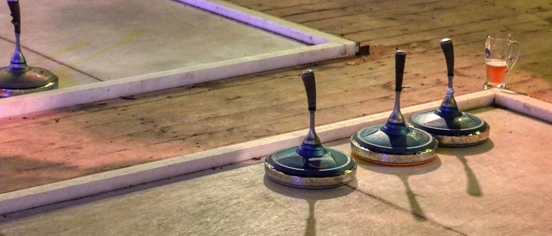 Drei Curlingsteine sind auf einem polierten Boden platziert, wobei Schatten auf der Oberfläche geworfen werden. Eine weiße Plattform befindet sich links.