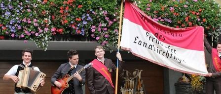 Three men dressed in suits and ties play musical instruments outside. One holds a guitar, another plays an accordion, and the third holds a flag.