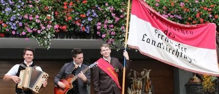 Three men in suits hold instruments and a flag, performing at an outdoor event. Behind them, a backdrop of flowers and a building is visible.