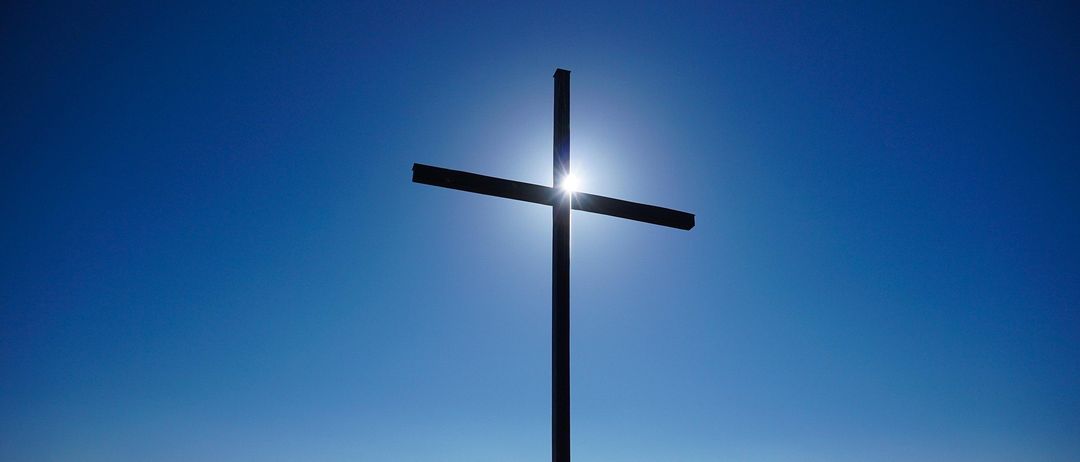 Ein großes Holzkreuz steht auf einem Hügel, mit der Sonne, die hell hinter ihm scheint, die das Kreuz beleuchtet und einen Schatten auf den Boden wirft.