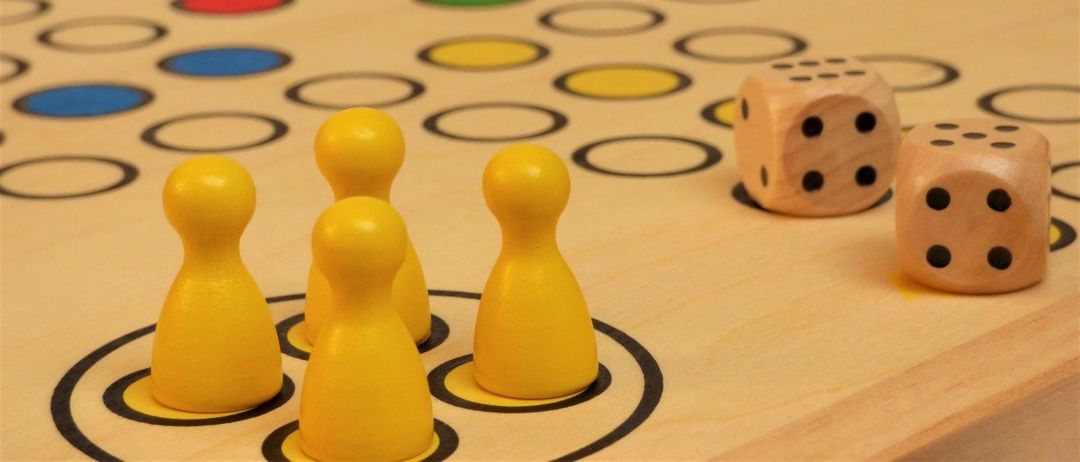 Ein Holzbrettspiel mit gelben und roten Spielfiguren, einschließlich Würfeln, ist mit einem Gittermuster aufgestellt. Die Spielfiguren sind um das Brett herum platziert.