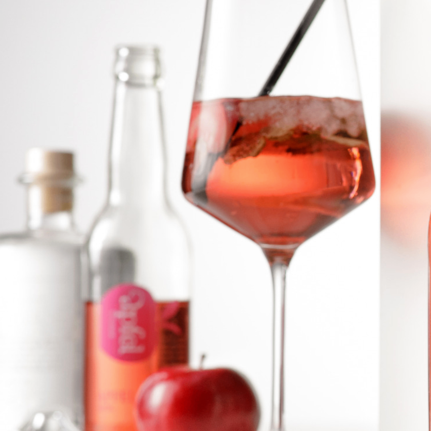 Ein Glas rosafarbener Cocktail, garniert mit einem schwarzen Strohhalm, steht neben einer Flasche Alkohol und einem roten Apfel vor weißem Hintergrund.