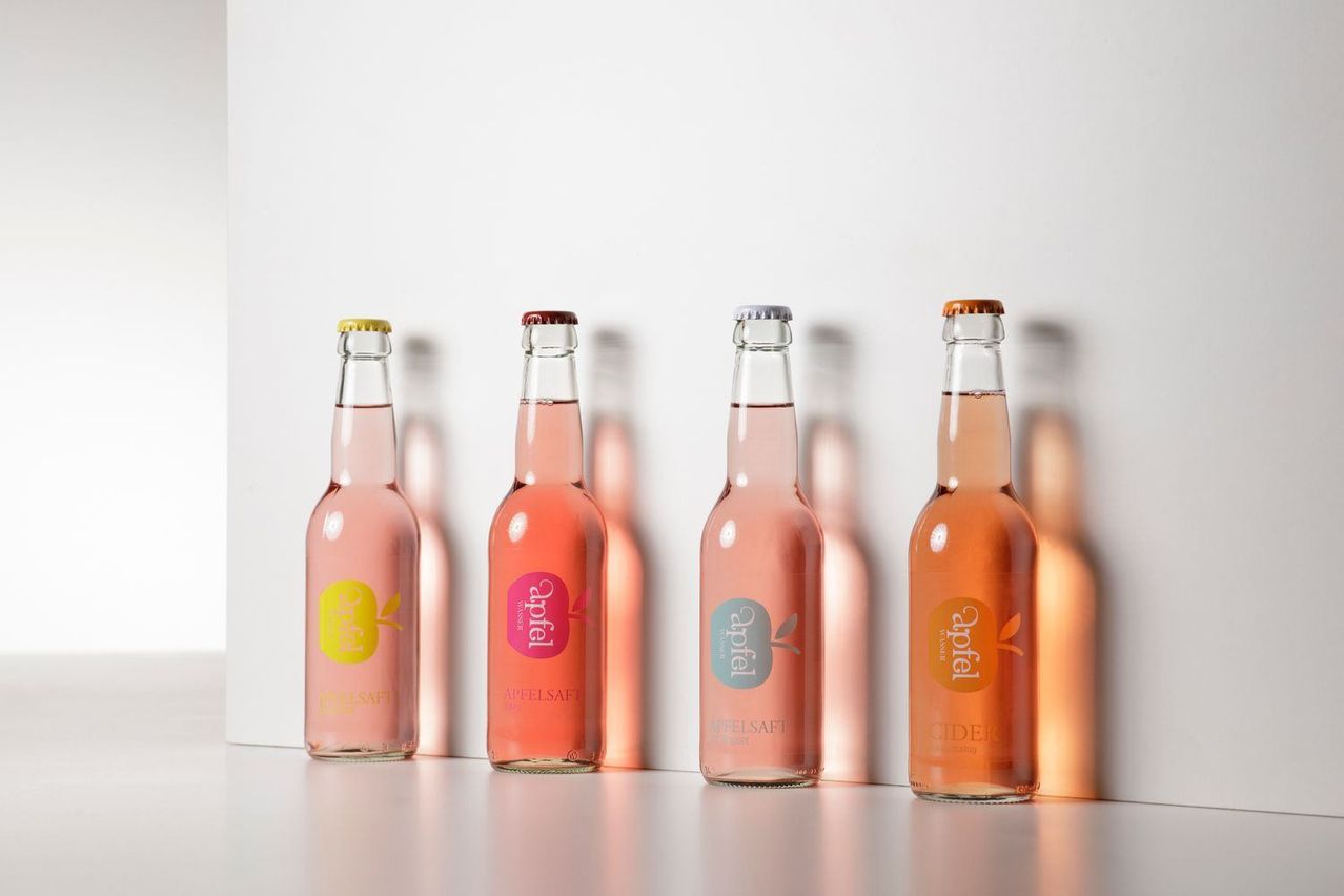Vier klare Flaschen mit rosa Flüssigkeit stehen in einer Reihe auf einer hellen Oberfläche. Sie sind mit Apfelsaft und Cider beschriftet. Der Hintergrund ist eine einfache weiße Wand.
