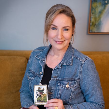 A woman in a denim jacket is sitting on a couch and smiling while holding a tarot card.