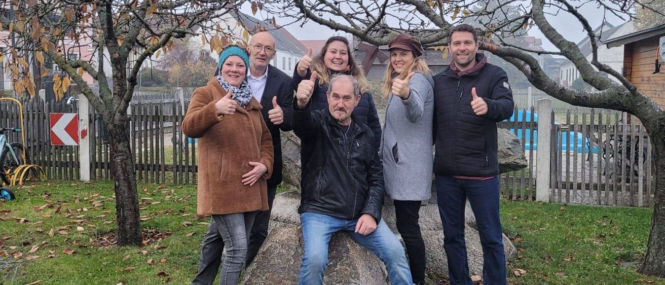 Fünf Menschen posieren für ein Foto in einem Park, zwei Frauen und drei Männer. Sie lächeln und geben Daumen hoch. Hinter ihnen steht ein großer Felsen und ein Baum. Das Gelände ist von einem Holzzaun umgeben.