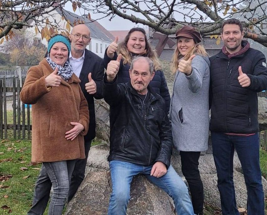 Fünf Menschen posieren für ein Foto in einem Park, zwei Frauen und drei Männer. Sie lächeln und geben Daumen hoch. Hinter ihnen steht ein großer Felsen und ein Baum. Das Gelände ist von einem Holzzaun umgeben.