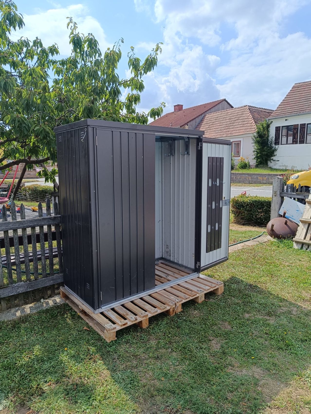 A black storage shed with an open door is placed on wooden pallets in a backyard. Behind it, a wooden fence and a tree are visible. In the background, there are houses with brown roofs.