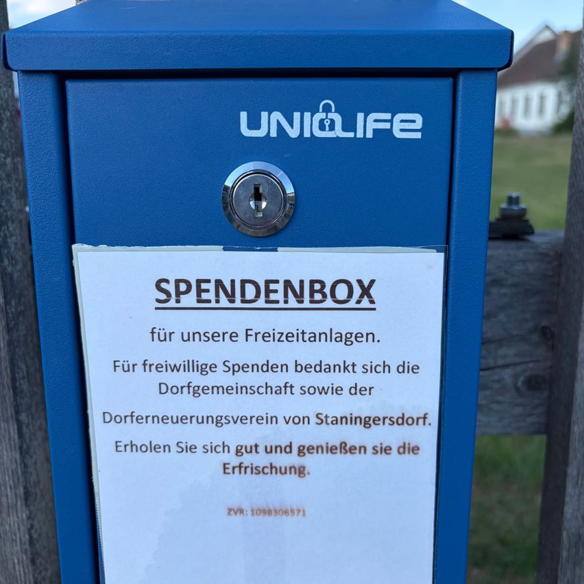 A blue Spendebox box with a lock, mounted on a wooden fence, has a white sign that reads 'Spendenbox' and 'for our leisure activities.'