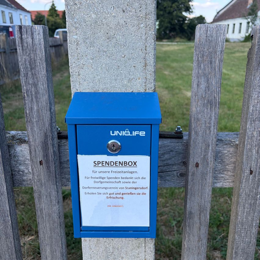 A blue donation box with the text 'Spendenbox' is mounted on a concrete post near a wooden fence. The box has a lock and a white label with German text. Behind the fence is a grassy field with a building in the distance.