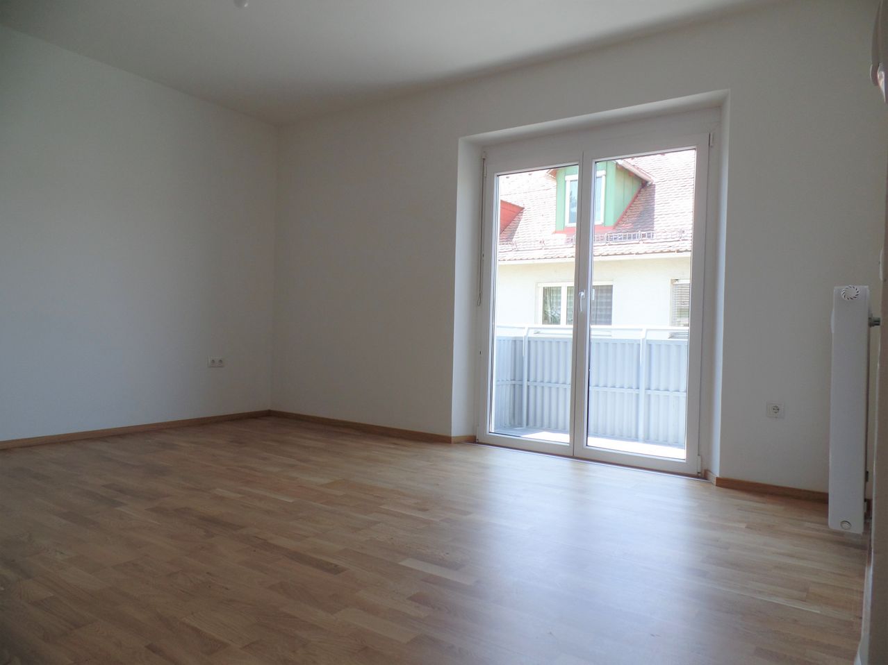 Ein leerer Raum mit Holzböden und weißen Wänden. Es gibt eine Schiebetür aus Glas, die zu einem Balkon mit Blick auf ein Haus mit einem grünen Dach führt.