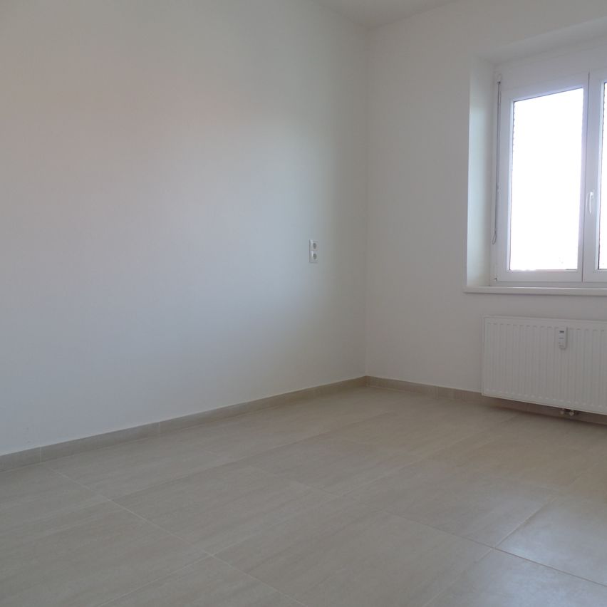 Ein leerer Raum mit weißen Wänden, gefliestem Boden, einem Fenster mit Jalousien und einem weißen Heizkörper an der Wand.