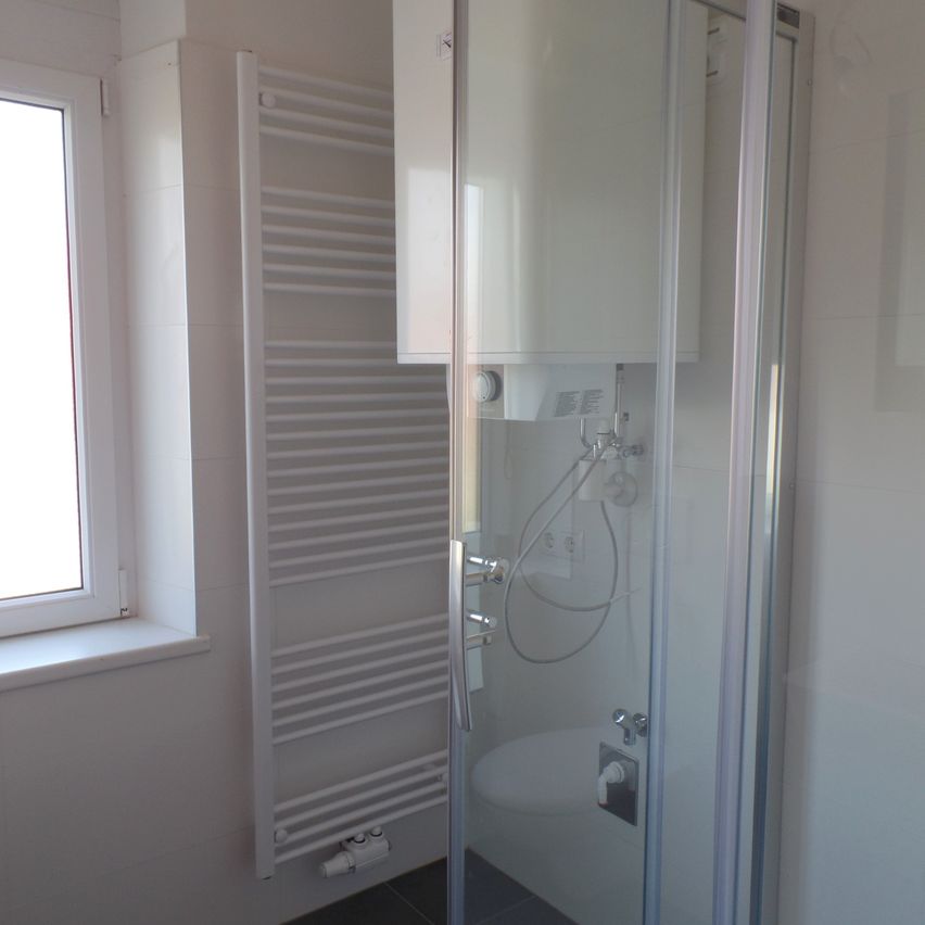 Ein modernes Badezimmer mit einer Glasdusche, weißen Fliesen, einem weißen Handtuchhalter und einem Fenster mit weißer Fensterbank.