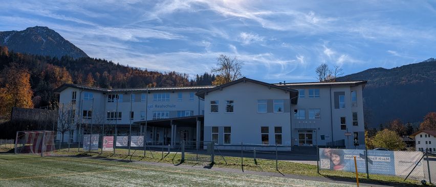 Ein weißes Gebäude mit vielen Fenstern steht vor einem Rasenfeld. Im Hintergrund befinden sich Berge und der Himmel ist blau mit Wolken.