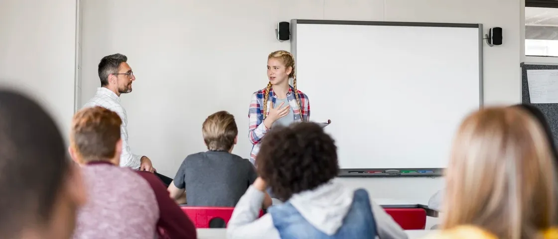 Ein Klassenzimmer mit einem jungen Mädchen, das vor einer Tafel präsentiert, während mehrere Schüler aufmerksam zuhören.