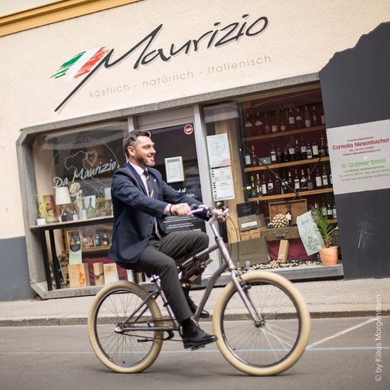 Ein Mann in einem Anzug fährt ein Fahrrad vor einem Geschäft namens Maurizio. Das Geschäft zeigt eine Vielzahl von Weinflaschen und anderen Produkten an.