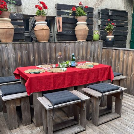 Eine Holzterrasse mit einem von einem roten Tischtuch bedeckten Tisch und vier Holzbänken. Dahinter befindet sich eine Holzwand mit mehreren großen Terrakotta-Töpfen mit roten Blumen.