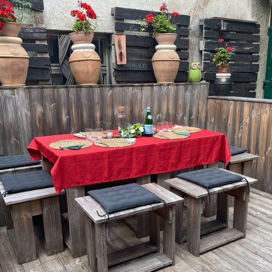 Eine Holzterrasse mit einem von einem roten Tischtuch bedeckten Tisch und vier Holzbänken. Dahinter befindet sich eine Holzwand mit mehreren großen Terrakotta-Töpfen mit roten Blumen.