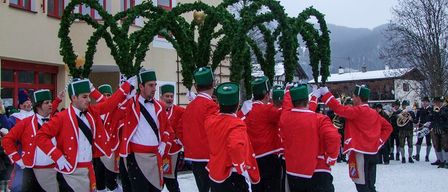 Eine Gruppe von Männern in roten Uniformen mit grünen Mützen marschiert im Schnee. Hinter ihnen steht ein Gebäude mit einem Kreuz oben.
