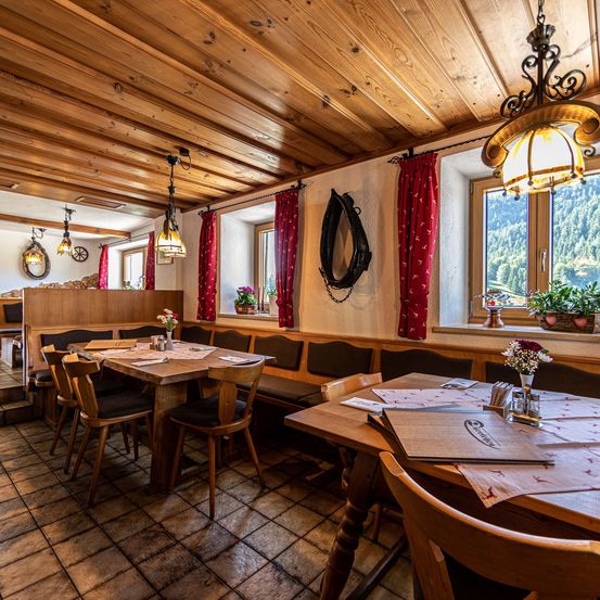 Ein leeres rustikales Restaurant mit Holzbänken, Tischen und gefliesten Böden. Ein Blick auf Berge und eine grüne Pflanze ist durch das Fenster zu sehen.