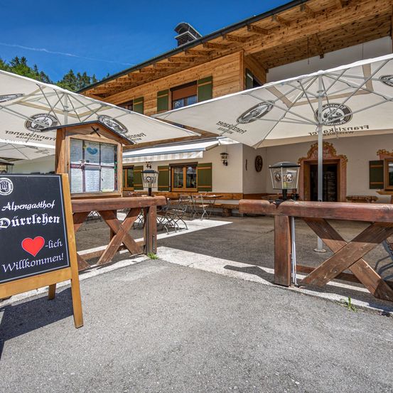 Ein Restaurant mit hölzerner Außenfassade hat ein Tafelschild vor sich. Es steht 'Alpengasthof Durrlehen - lich Willkommen!' Es gibt Tische im Freien mit Sonnenschirmen.