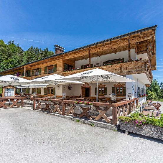 Ein Chalet-Restaurant aus Holz mit Balkon und mehreren weißen Sonnenschirmen. Die Vorderseite hat einen Zaun mit Blumenkästen. Bäume sind im Hintergrund.