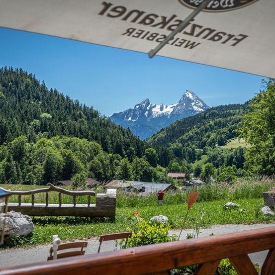 Eine Holzbank steht in der Nähe einer Straße mit Blick auf eine Bergkette. Ein großer Regenschirm ist darüber und verdeckt teilweise die Sicht.