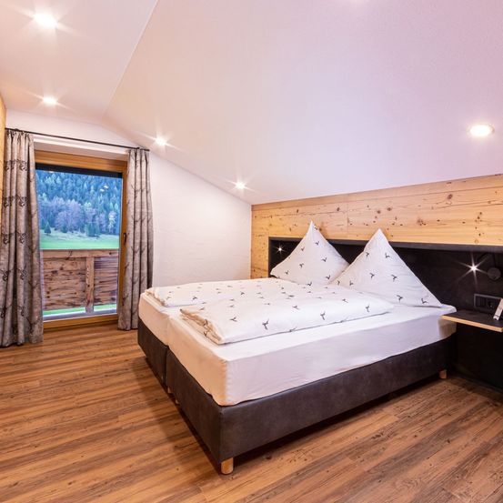 Ein Schlafzimmer mit Holzwänden, einem Bett mit weißer Bettwäsche und zwei weißen Kissen mit Vogeldesign. Es gibt ein Fenster mit Vorhängen, einen Balkon und eine Aussicht auf einen Berg.