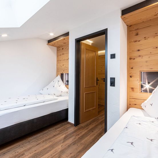 Ein Schlafzimmer mit zwei Betten, Holzwänden und Bodenbelag. Jedes Bett hat eine weiße Bettwäsche mit Vogelmuster und zwei weißen Kissen. Eine Tür mit hölzernerahmen ist offen.