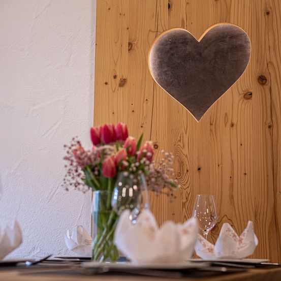 Eine Holzwand mit ausgeschnittenem Herz zeigt ein Herz im Inneren. Eine Vase mit roten Tulpen steht auf einem Tisch mit Weingläsern, gefalteten Servietten und Tellern.