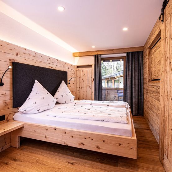 Ein Schlafzimmer mit Holzwänden und -böden, mit einem Doppelbett mit gemusterten Kissen, einem Nachttisch und einem Fenster mit Vorhängen.