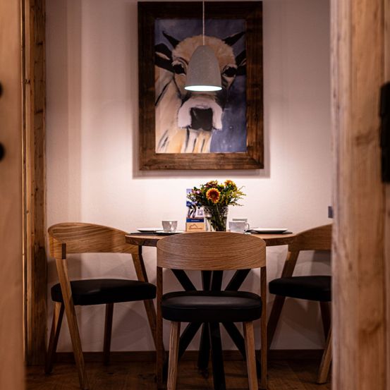 Ein gemütlicher Essbereich mit einem Holztisch, vier Stühlen und einem Kuhbild an der Wand. Eine Vase mit Blumen steht auf dem Tisch, und eine Lampe hängt von der Decke.