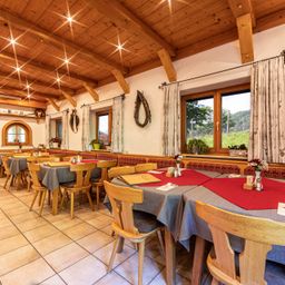 Ein Restaurant mit Holzdecke, Tischen, Stühlen und großen Fenstern. Gepflanzte Pflanzen und dekorative Gegenstände schmücken die Wände. Der Raum ist in warmem, natürlichem Licht gebadet.