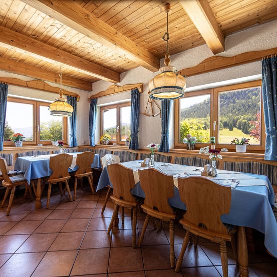 Ein leerer Essraum mit blauen Tischdecken, Holzstühlen und Fenstern mit blauen Vorhängen. Der Raum hat Holzbalken und einen Blick auf die Berge.