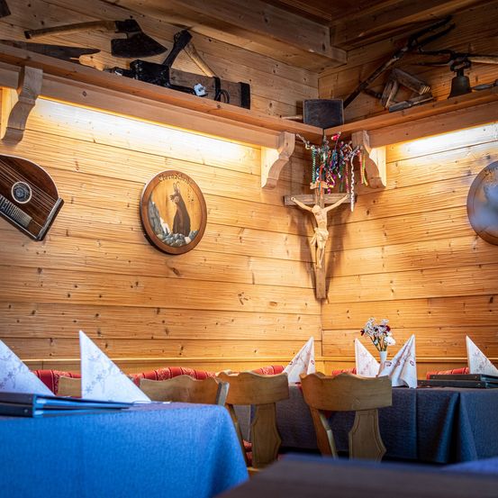 Ein hölzernes Interieur mit Tischen und Stühlen, ein Tisch hat blaues Tuch, ein Kreuz und Blumen. Über den Tischen befinden sich Regale mit verschiedenen Gegenständen, einschließlich einer Glocke. Die Wände sind aus Holz mit einer aufgeleuchteten Lampe.