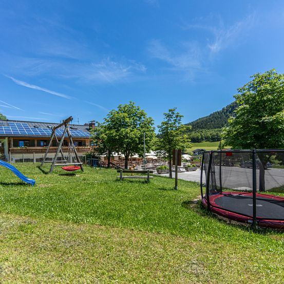 Ein Spielplatz mit einem blauen Rutschen, Schaukeln und einem Trampolin unter einem sonnigen Himmel. Dahinter ein Gebäude mit Solarpanelen und einem Hügel mit Bäumen.