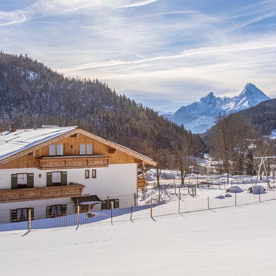 Ein Chalet steht in einer verschneiten Landschaft mit einem Zaun, Bergen im Hintergrund und einem klaren Himmel.
